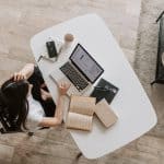 From above of young woman with long dark hair in casual clothes working at table and browsing netbook while sitting in modern workplace and touching hair